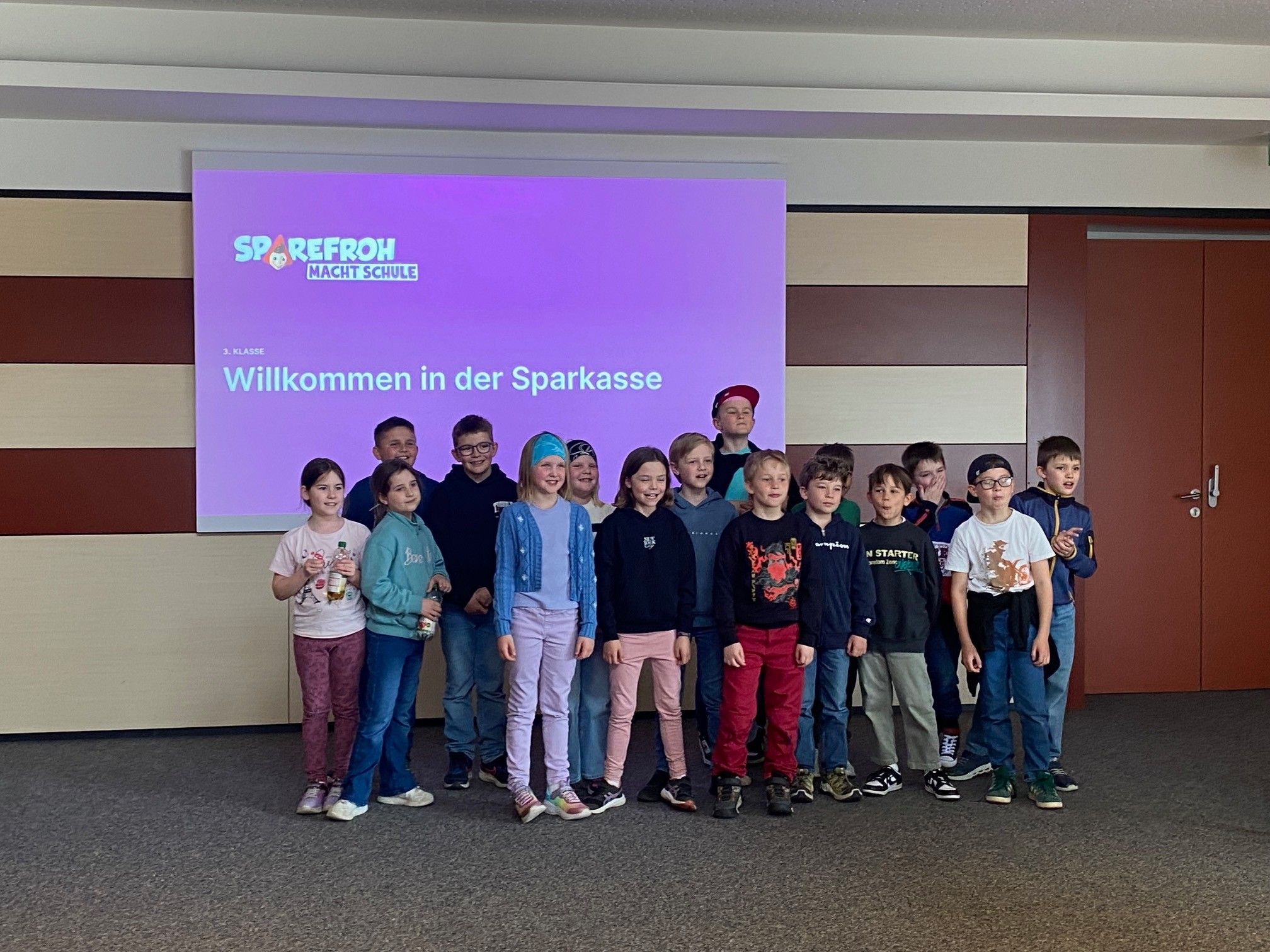 Eine Gruppe von Kindern und Erwachsenen steht vor einer Projektionsleinwand mit einer Präsentation über Sparfrosch Macht Schule.