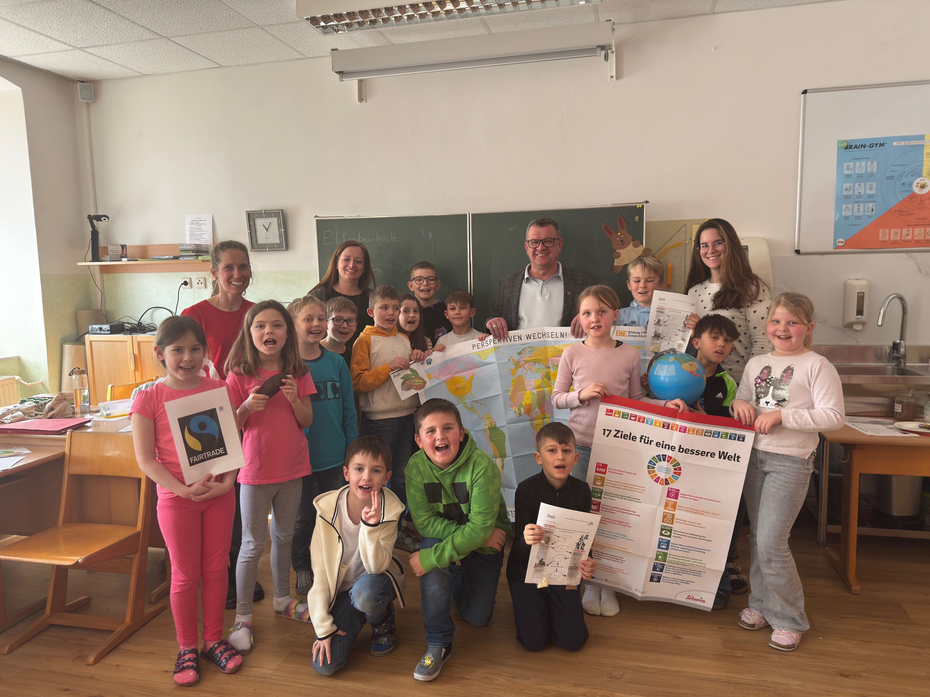 Eine Gruppe von Kindern und Erwachsenen posiert für ein Foto im Klassenzimmer, hält eine Karte und einen Globus. Sie alle lächeln und scheinen an einer Lernaktivität beteiligt zu sein.