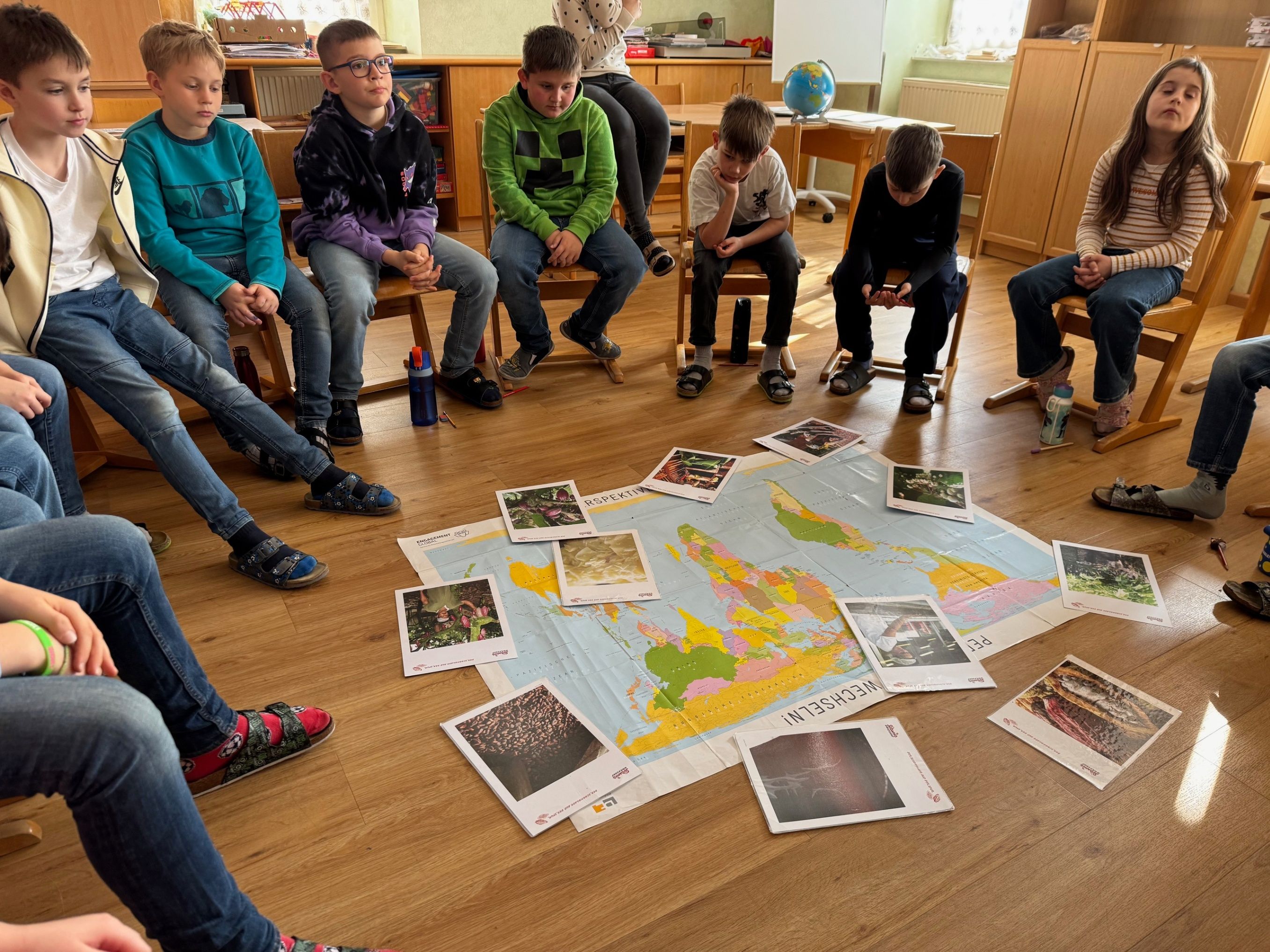 Eine Gruppe von Kindern sitzt auf Stühlen um eine große Weltkarte auf dem Boden herum und betrachtet Bilder und diskutiert.
