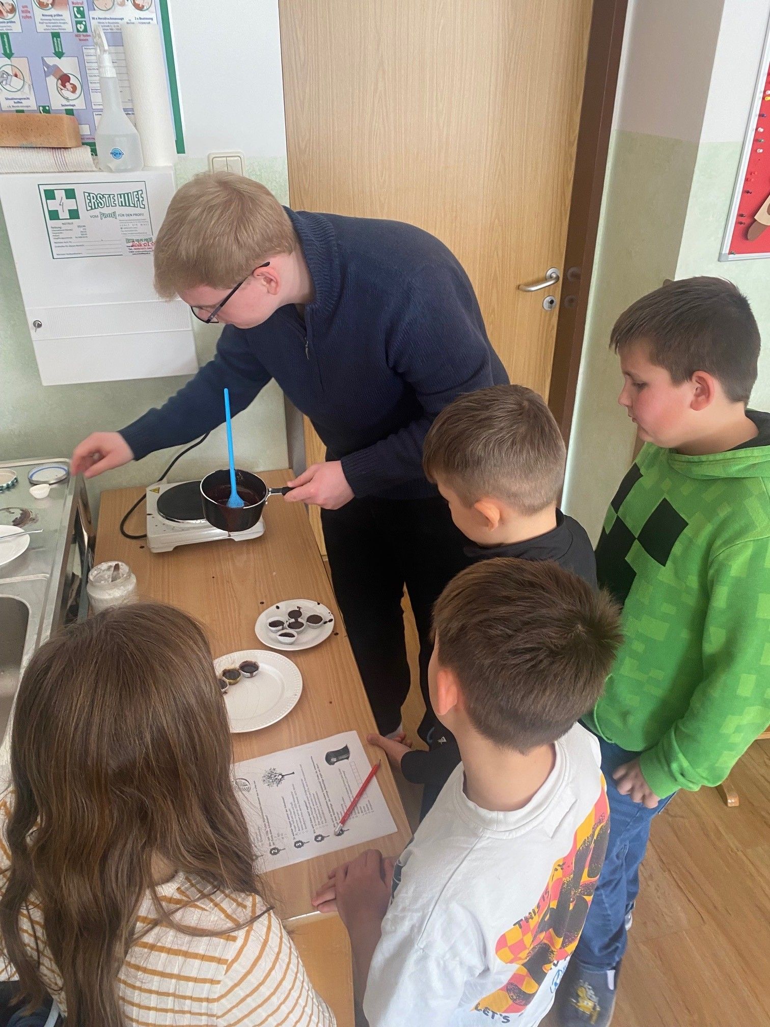 Ein Mann in einem blauen Pullover kocht auf einem heißen Teller, während eine Gruppe von Kindern zuschaut. Die Kinder stehen um einen Tisch mit Tellern voller Essen.
