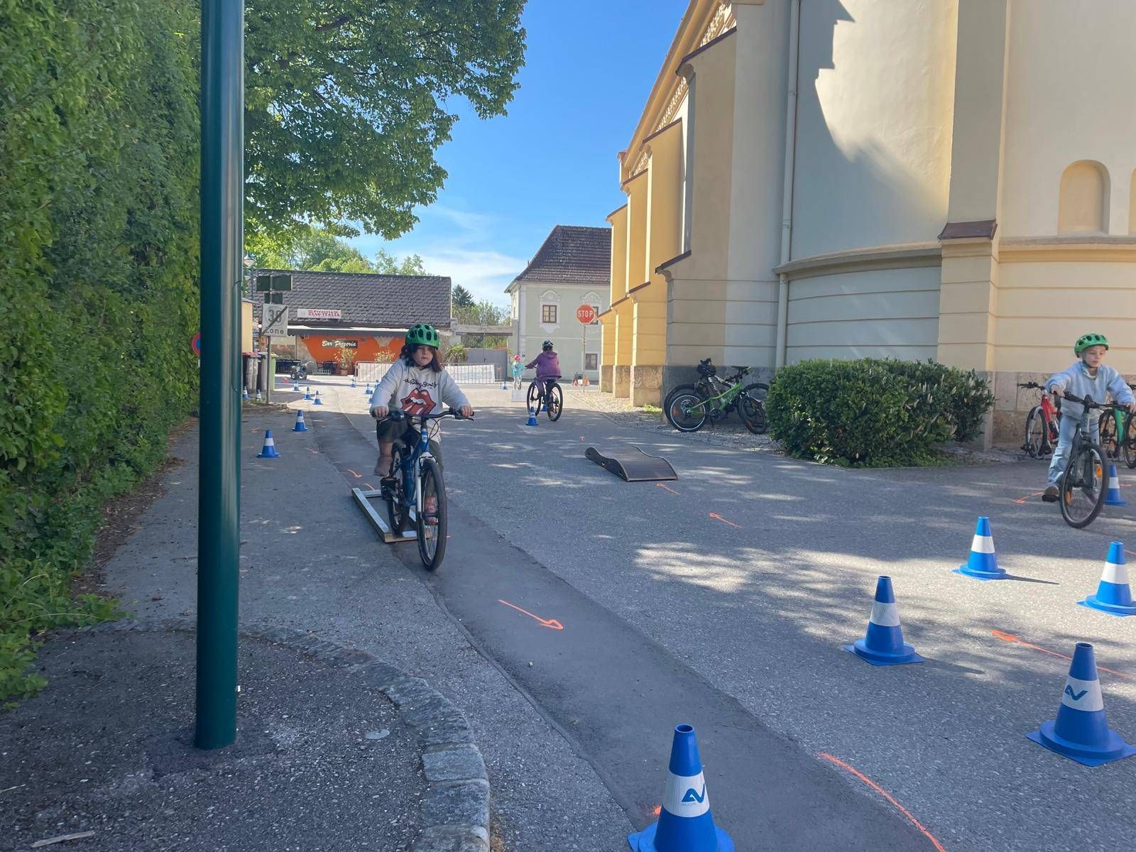 Ein Kind fährt auf einem Fahrrad auf einer Rampe in einem Parkplatz, mit Verkehrskegel und Stoppschild im Hintergrund.
