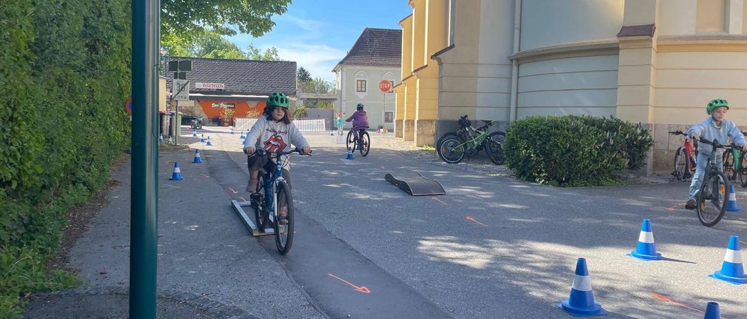 Ein Kind fährt auf einer Rampe auf einem Fahrrad in einer Straße, umgeben von Verkehrskegel und geparkten Fahrrädern.