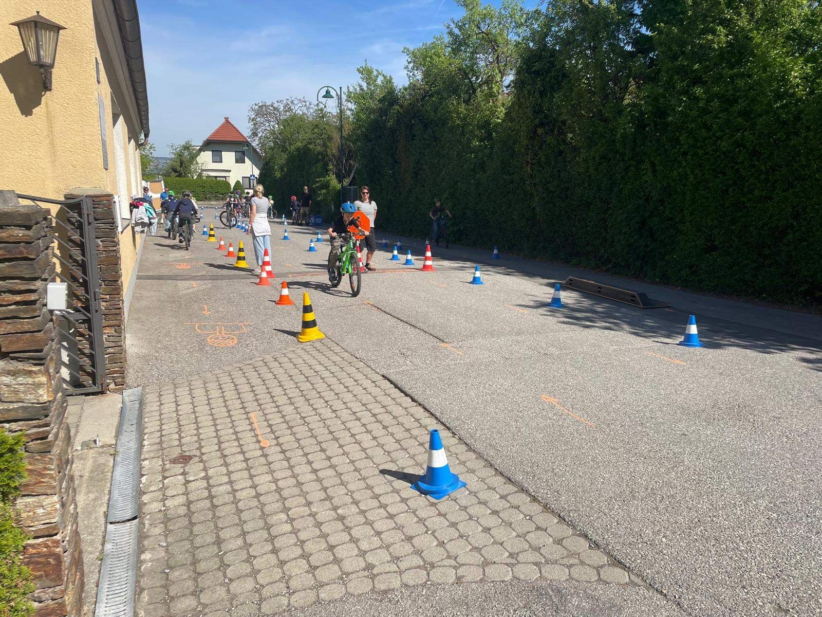 Kinder üben das Radfahren auf einer gepflasterten Straße, umgeben von orangefarbenen und blauen Kegel. Ein paar Erwachsene beaufsichtigen.