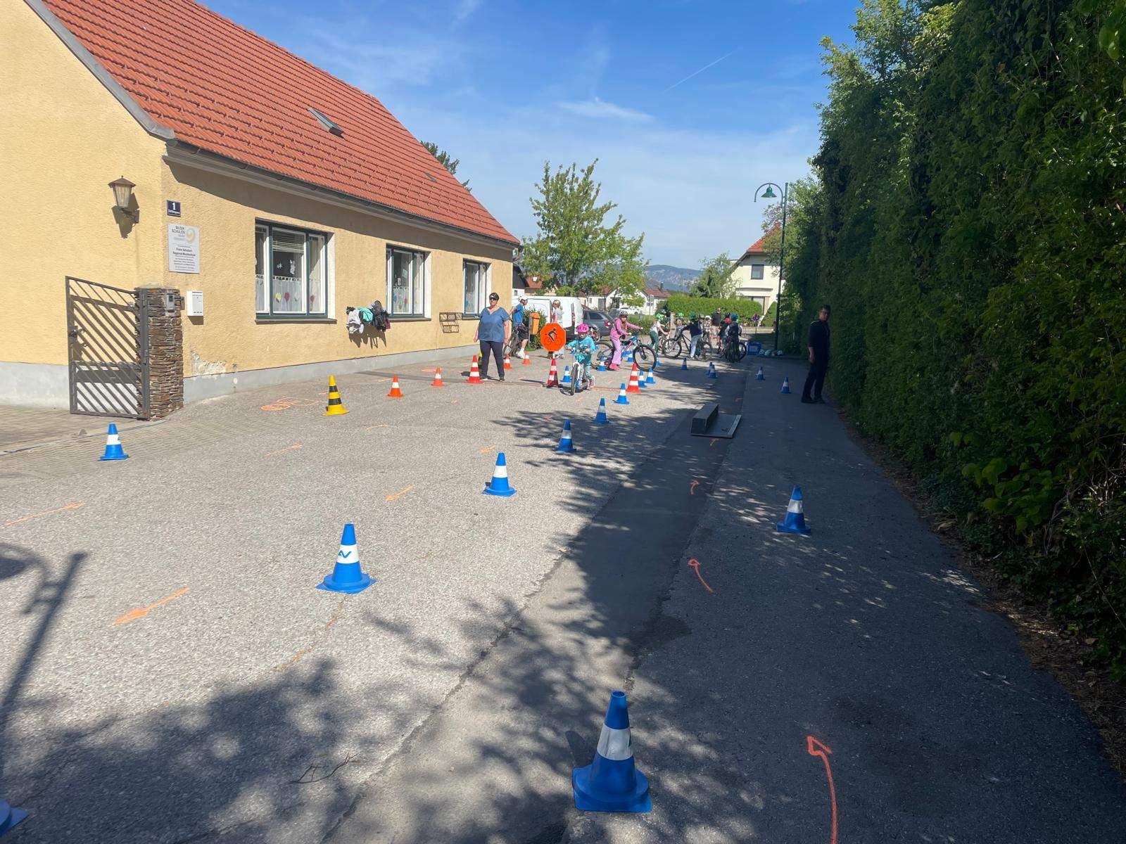 Eine Gruppe von Kindern übt das Radfahren auf einer Straße mit Verkehrskegeln und einer Frau in der Nähe.