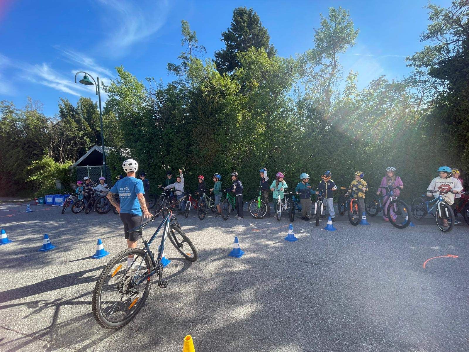Eine Gruppe von Kindern steht mit ihren Fahrrädern in einer Reihe, trägt Helme und steht vor einer Reihe von Hütchen. Ein Mann in einem blauen Shirt steht neben einem Fahrrad.