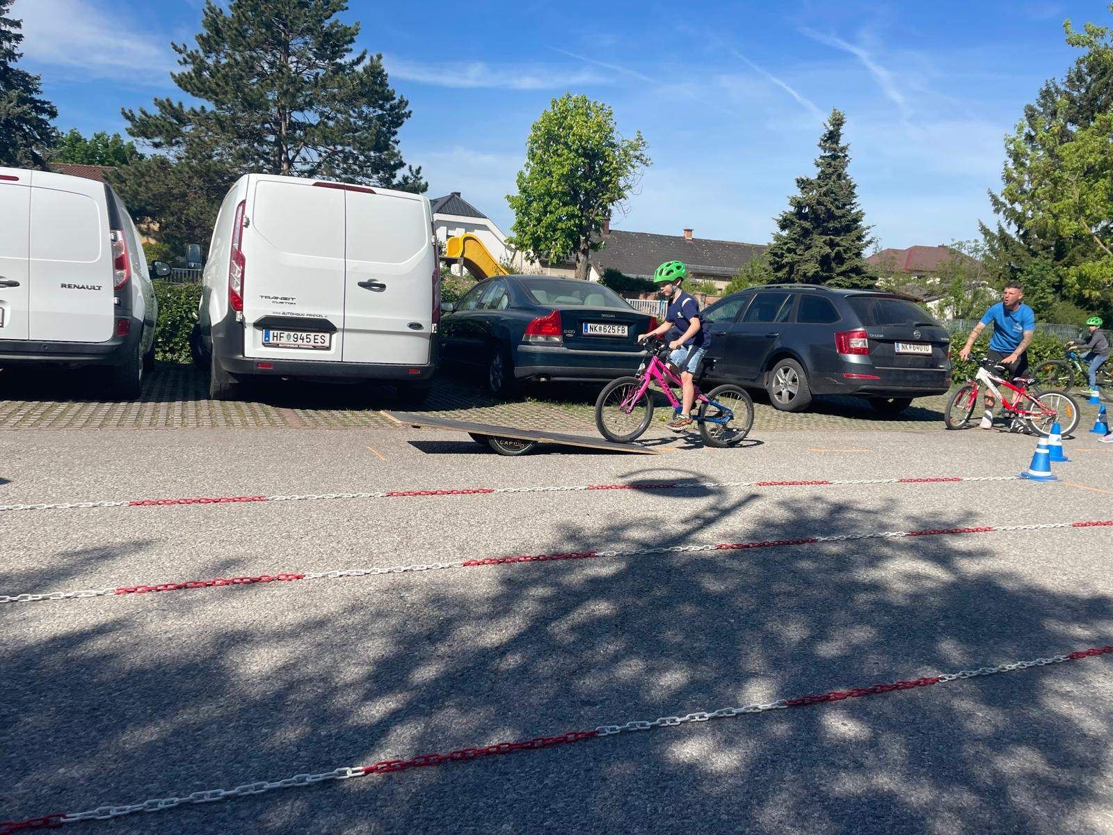 Ein Kind fährt ein pinkes Fahrrad auf eine Rampe in einem Parkplatz, mit einem weißen Lieferwagen und zwei Autos im Hintergrund.