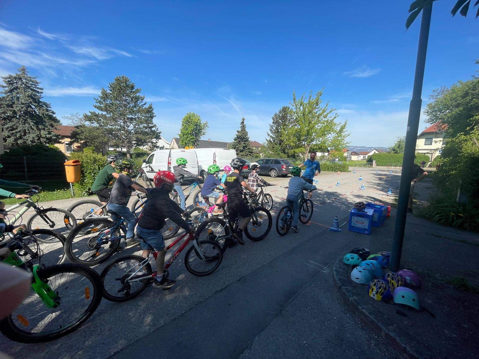 Eine Gruppe von Radfahrern, die Helme tragen, steht auf einer Straße, wahrscheinlich für eine Radveranstaltung. Ein Mann in einem blauen Shirt steht hinter ihnen. Es sind mehrere Fahrräder geparkt, zusammen mit einigen orangefarbenen Kegeln. In der Ferne sind ein weißer Lieferwagen und ein Auto geparkt, in der Nähe einiger Bäume.