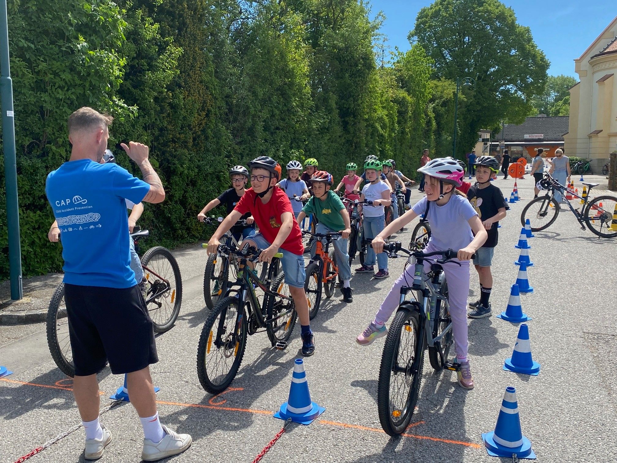 Eine Gruppe von Kindern mit Helmen fährt in einer Reihe auf Fahrrädern, begleitet von einem Erwachsenen in einem blauen T-Shirt. Sie sind von blauen Kegel und Bäumen umgeben.
