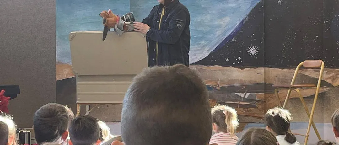 Ein Mann in Pilotenuniform steht vor einem Klassenzimmer, hält eine Puppe und hat ein Wandbild im Hintergrund. Kinder sitzen vorne und schauen aufmerksam zu.