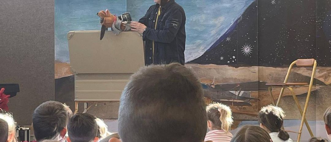 Ein Mann in Pilotenuniform steht vor einem Klassenzimmer, hält eine Puppe und hat ein Wandbild im Hintergrund. Kinder sitzen vorne und schauen aufmerksam zu.