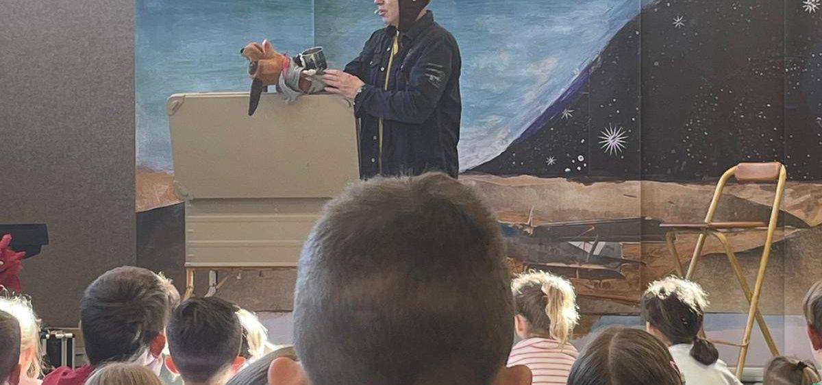 Ein Mann in Pilotenuniform steht vor einem Klassenzimmer, hält eine Puppe und hat ein Wandbild im Hintergrund. Kinder sitzen vorne und schauen aufmerksam zu.