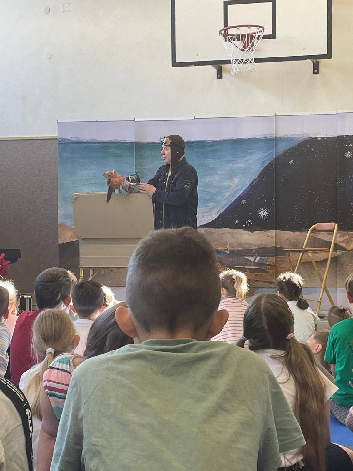 Ein Mann in Pilotenuniform steht vor einer großen Wandmalerei und hält eine Puppe, während eine Klasse von Kindern zuschaut.