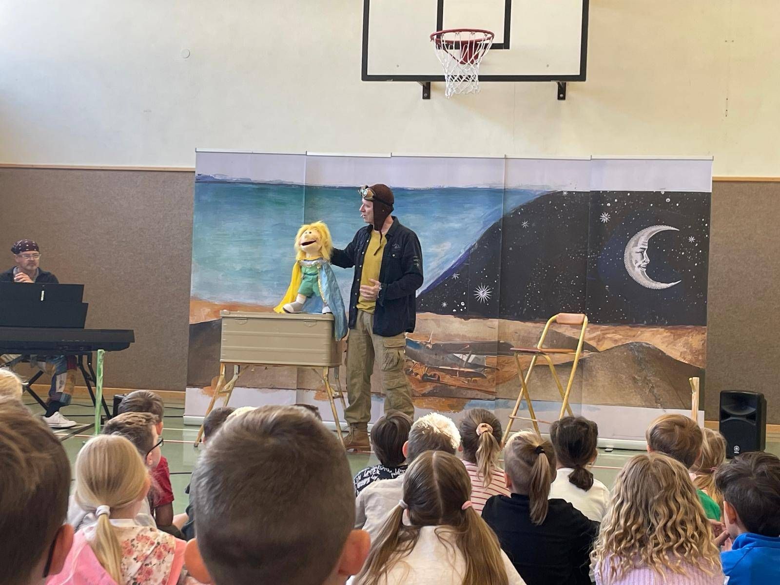 Ein Mann führt eine Puppenshow vor einem großen bemalten Hintergrund in einer Schulsporthalle auf.