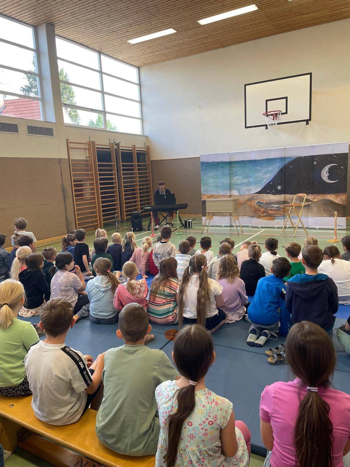 Eine Gruppe von Kindern ist in einer Turnhalle versammelt und hört einer Person zu, die Klavier spielt. Der Raum hat große Fenster und eine Wandmalerei.