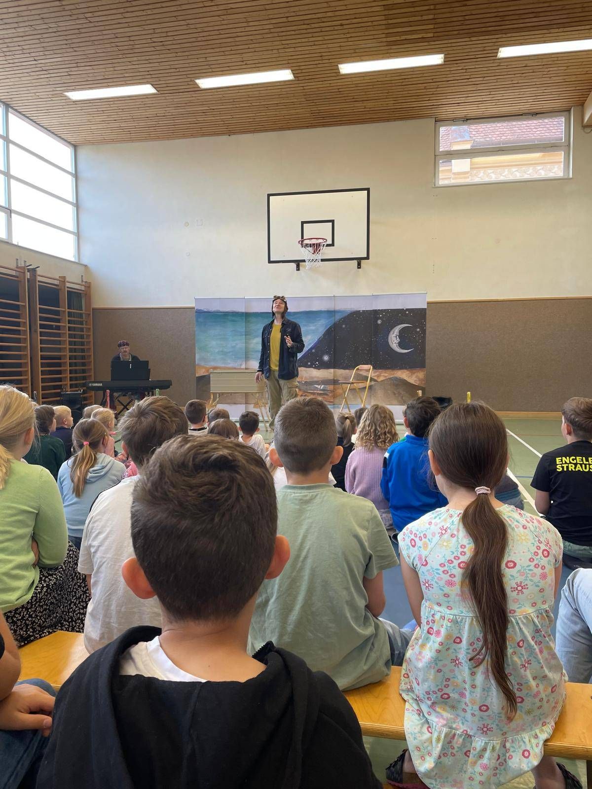 Eine Person steht auf der Bühne in einer Turnhalle und tritt vor einer Gruppe von Kindern auf. Im Hintergrund ist eine Kulisse mit Nachthimmel und Ozean zu sehen.