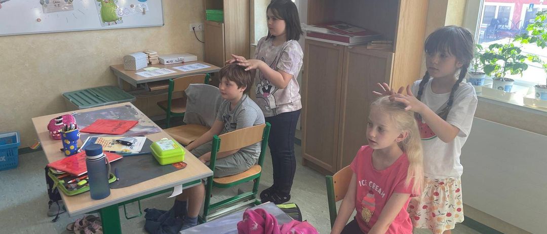 Drei Kinder sind in einem Klassenzimmer, eines lässt sich die Haare von einem anderen frisieren, während das dritte zuschaut. Ein Schreibtisch ist mit Büchern und Utensilien übersät.