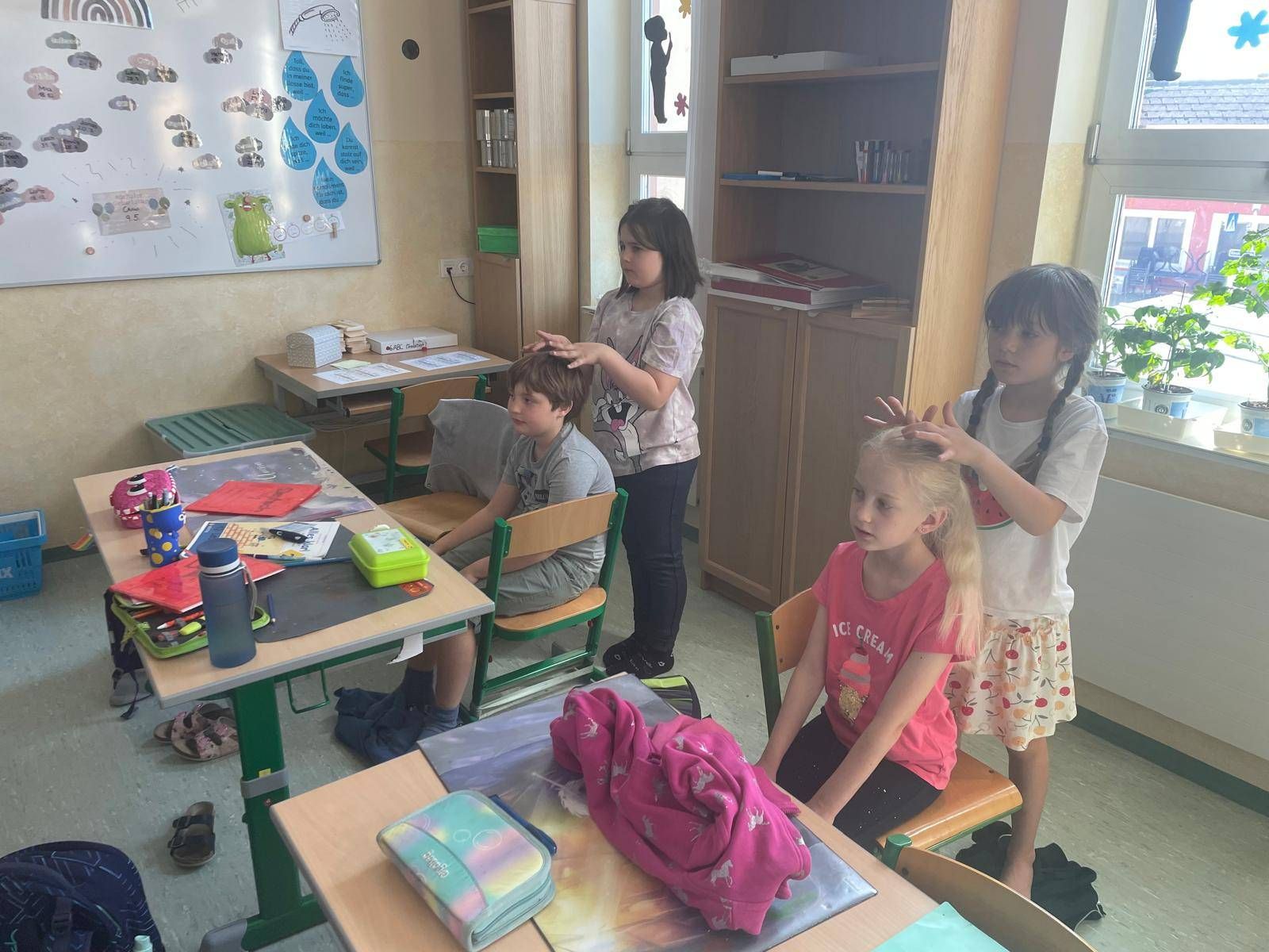Drei Kinder in einem Klassenzimmer, ein Mädchen bürstet die Haare eines anderen Mädchens, und ein Junge sitzt auf einem Stuhl.