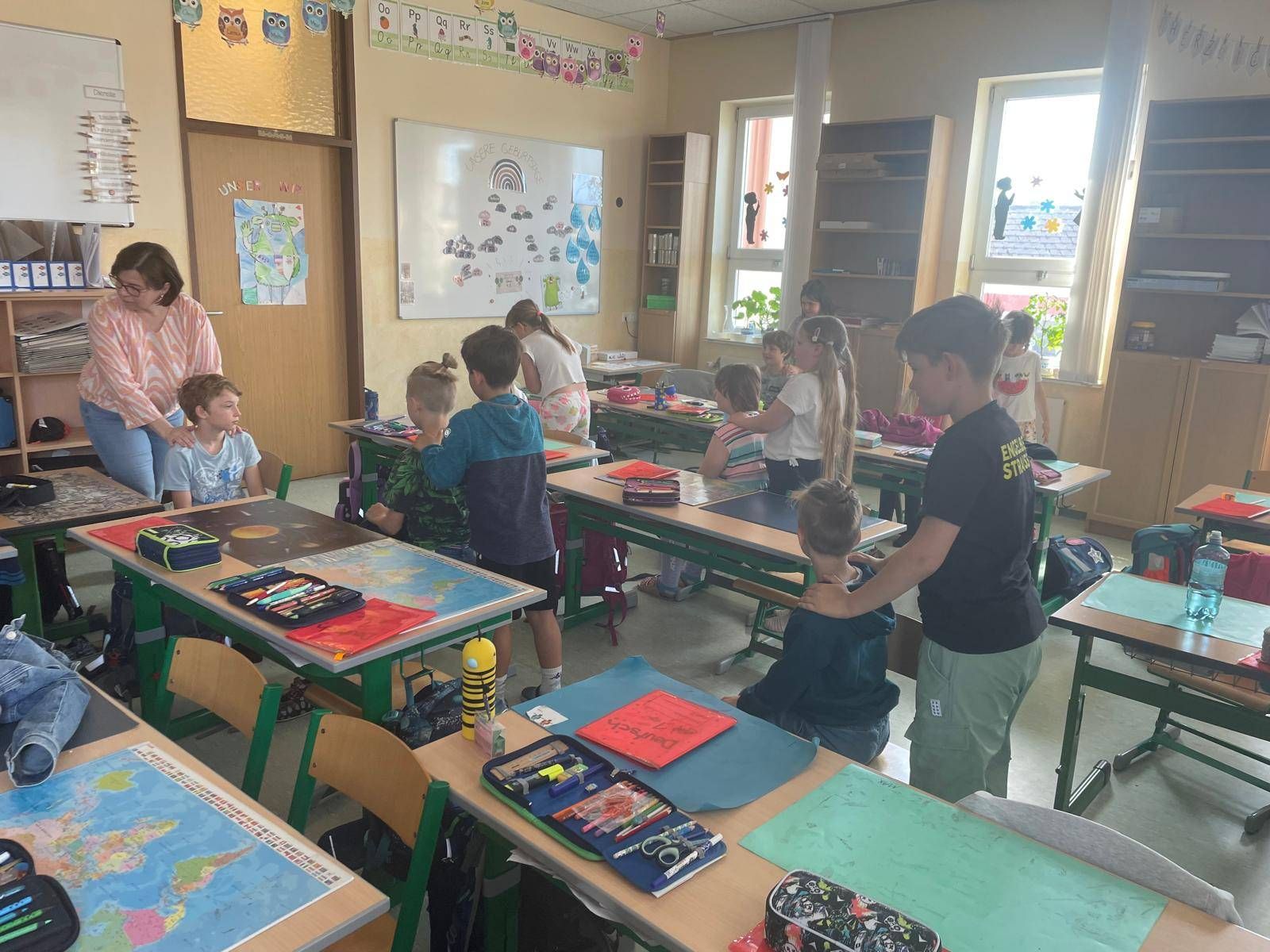 Ein Klassenzimmer mit Kindern, die Kunstprojekte bearbeiten, eine Lehrerin hilft einem Jungen und eine Tafel mit Zeichnungen.