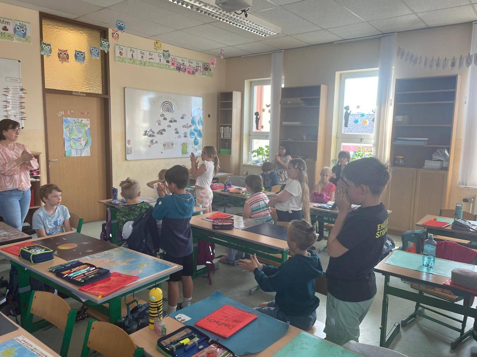 Ein Klassenzimmer mit Kindern, die an einer Aktivität teilnehmen, einige stehend und andere sitzend an Schreibtischen, mit einer Lehrerin im Hintergrund.