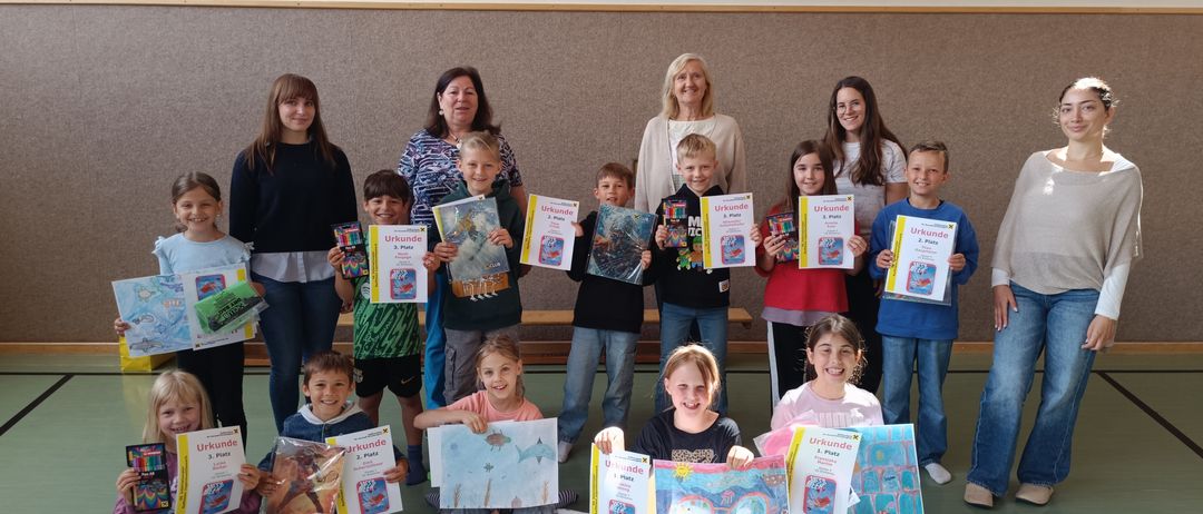 Eine Gruppe von Kindern und Erwachsenen posiert für ein Foto in einer Turnhalle, hält Urkunden und Bücher in den Händen. Die Kinder sitzen auf dem Boden, während die Erwachsenen hinter ihnen stehen. Der Raum hat einen Basketballkorb und eine Wand mit einem Basketballnetz.