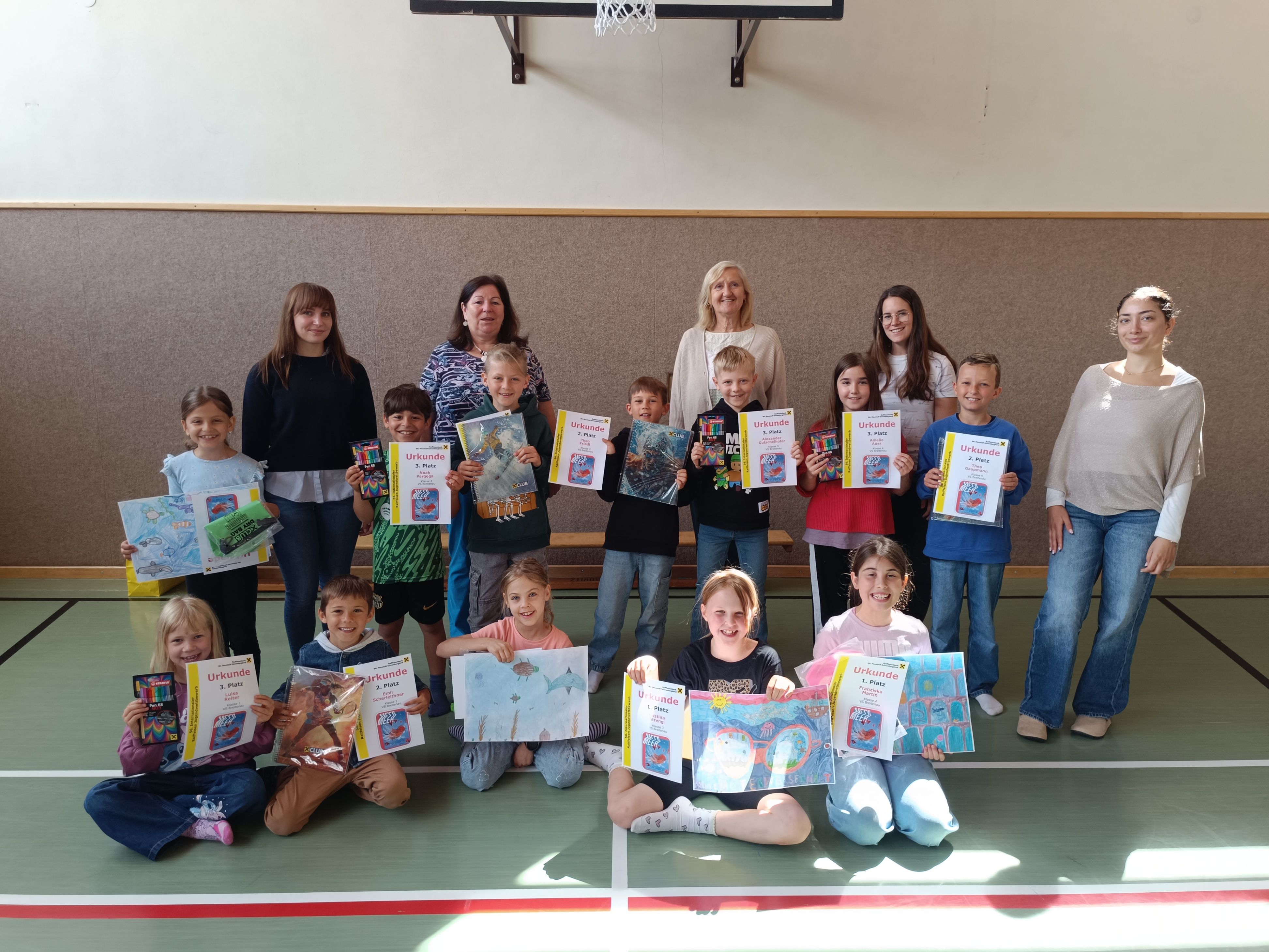 Eine Gruppe von Kindern und Erwachsenen posiert für ein Foto in einer Turnhalle, hält Urkunden und Bücher in den Händen. Einige sitzen auf dem Boden, andere stehen. Ein Basketballkorb ist im Hintergrund zu sehen.