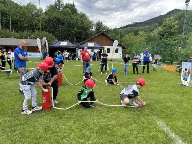 Kinder in Helmen nehmen an einem Tauziehen auf einem Rasenfeld teil, während Erwachsene beaufsichtigen. Zelte und ein Gebäude sind im Hintergrund zu sehen.