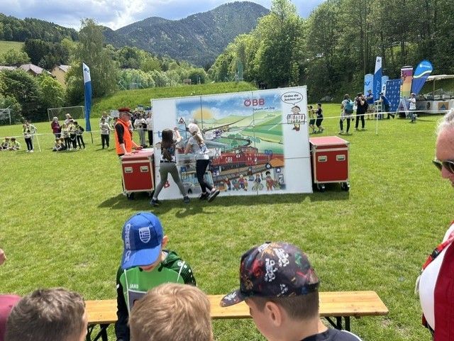 Eine Gruppe von Kindern und Erwachsenen versammelt sich um ein großes Brett mit einem Zugthema, möglicherweise an einer Veranstaltung auf einem Rasenfeld mit Bergen im Hintergrund.