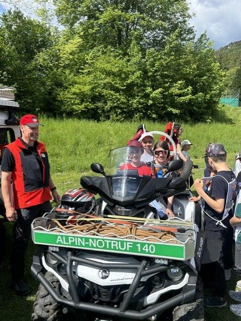 Eine Gruppe von Menschen versammelt sich um ein Geländefahrzeug, wobei eine Person auf dem Fahrersitz sitzt. Sie tragen alle Mützen und Sonnenbrillen, und einige haben Rucksäcke. Das Fahrzeug hat oben eine grüne Box mit der Aufschrift 'ALPIN-NOTRUF 140'. Dahinter befindet sich eine Grasfläche mit Bäumen und einem Berg in der Ferne.