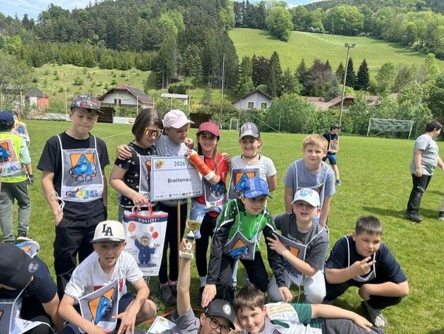 Eine Gruppe von Kindern auf einem Rasenfeld, einige tragen Hüte und halten Pokale, mit Häusern und Hügeln im Hintergrund.