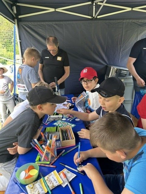 Eine Gruppe von Kindern sitzt unter einem Zelt um einen Tisch herum und malt mit Markern und Bleistiften. Ein Erwachsener steht hinter ihnen, möglicherweise zur Aufsicht.