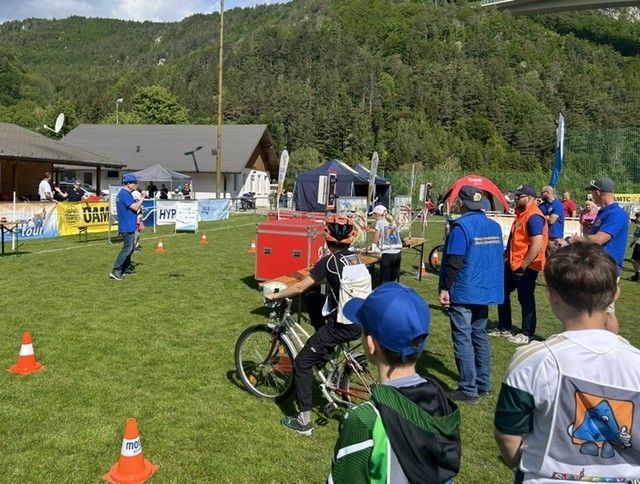 Eine Gruppe von Menschen versammelt sich auf einem Rasenfeld, möglicherweise für eine Radveranstaltung. Ein Mann fährt ein Fahrrad, während andere zuschauen. Es gibt Zelte, Verkehrskegel und Banner im Bereich. In der Ferne gibt es Berge und Bäume.
