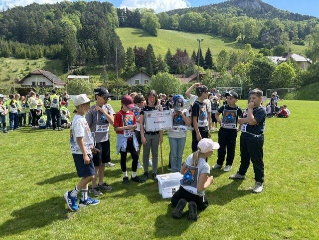 Eine Gruppe von Menschen, einschließlich Kindern, steht auf einem Feld mit Bergen im Hintergrund. Einige halten ein Banner und eine Person kniet.