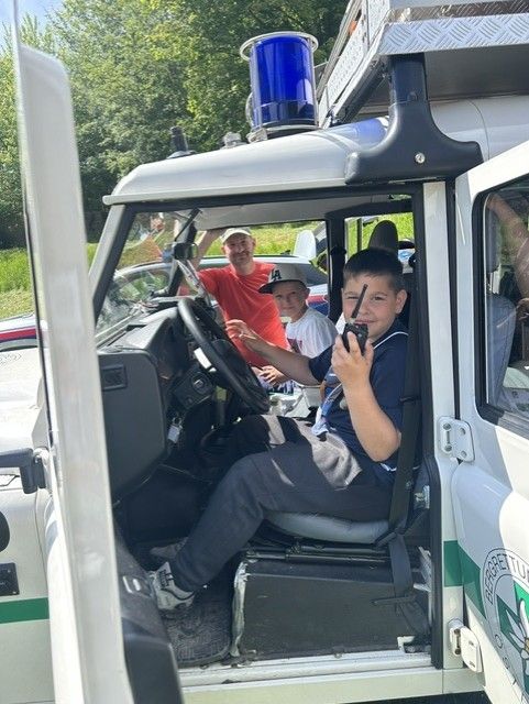 Ein Junge in einem blauen Shirt sitzt auf dem Fahrersitz eines Fahrzeugs und hält ein Walkie-Talkie, während zwei Männer in Hüten zusehen.