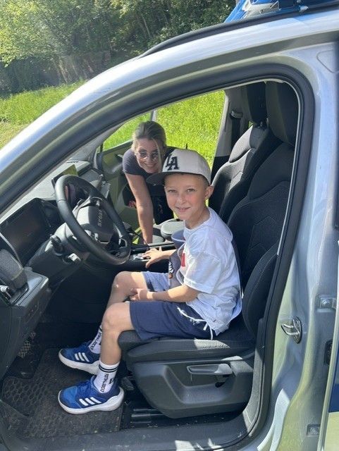 Ein junger Junge sitzt im Fahrersitz eines Autos, trägt eine Baseballkappe und Turnschuhe. Eine Frau steht hinter ihm und repariert möglicherweise etwas. Das Auto ist auf einem Grasfeld geparkt.