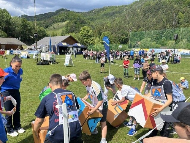 Eine Gruppe von Kindern und Erwachsenen nimmt auf einem Rasenfeld an einem Spiel teil, mit Bergen im Hintergrund. Sie halten Kisten und scheinen an einem Rennen beteiligt zu sein.