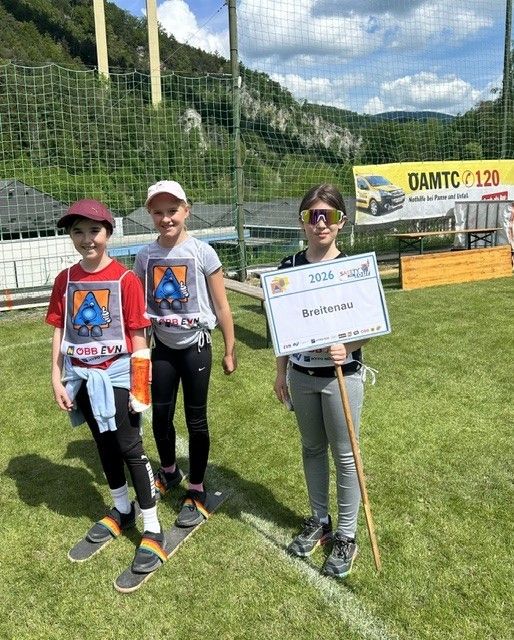 Drei junge Mädchen stehen auf einem Rasenfeld, tragen Turnschuhe und halten ein Schild mit der Aufschrift 'Breitenau 2026'. Das Mädchen rechts trägt eine Sonnenbrille und einen Gürtel. Dahinter steht ein Zaun mit einem Banner.