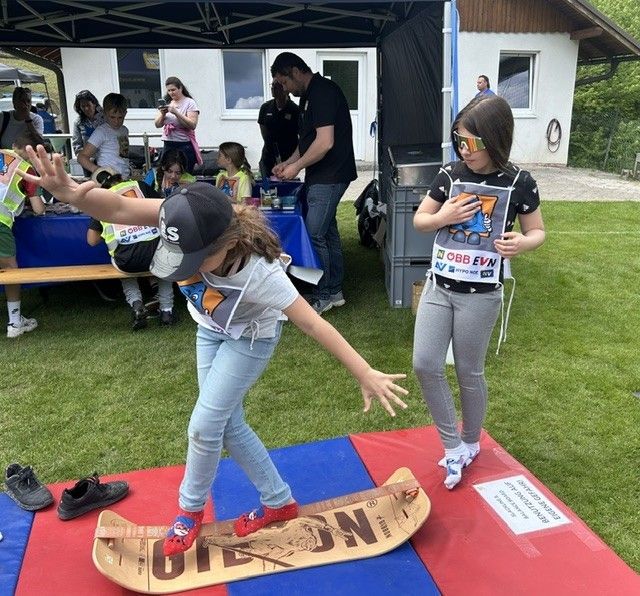 Zwei Mädchen stehen auf einem Snowboard, eines balanciert, während das andere zuschaut. Sie befinden sich in einem Grasbereich mit einem Gebäude und Menschen im Hintergrund.