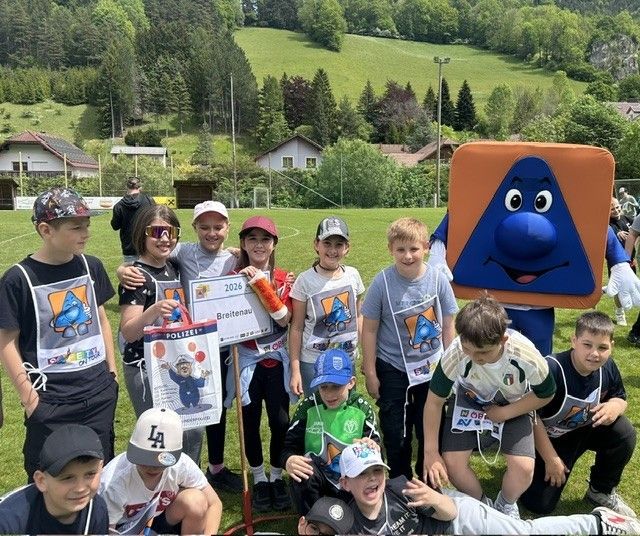 Eine Gruppe von Kindern in passenden T-Shirts mit einem Maskottchen im Hintergrund, posiert für ein Foto auf einem Rasenfeld.