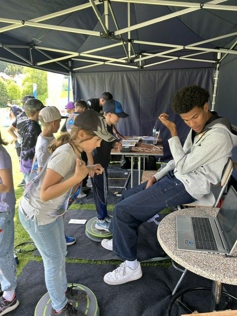 Eine Gruppe von Kindern und Erwachsenen versammelt sich unter einem Zelt. Ein junges Mädchen steht auf einem Balancebrett, während ein Junge auf einem Stuhl mit Laptop sitzt.