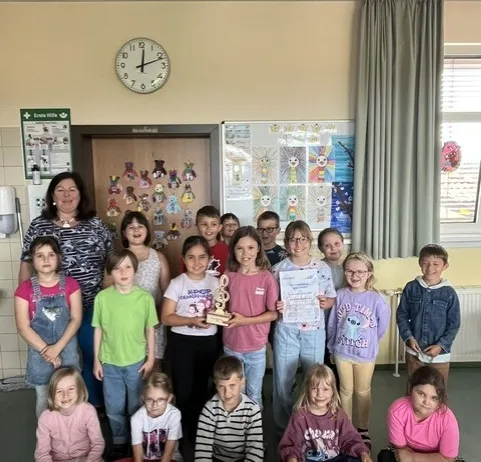 Eine Gruppe von Kindern und einer erwachsenen Frau posiert für ein Foto in einem Klassenzimmer. Sie halten einen Pokal und Urkunden. Das Klassenzimmer hat eine Uhr, eine Tafel und einen Vorhang.