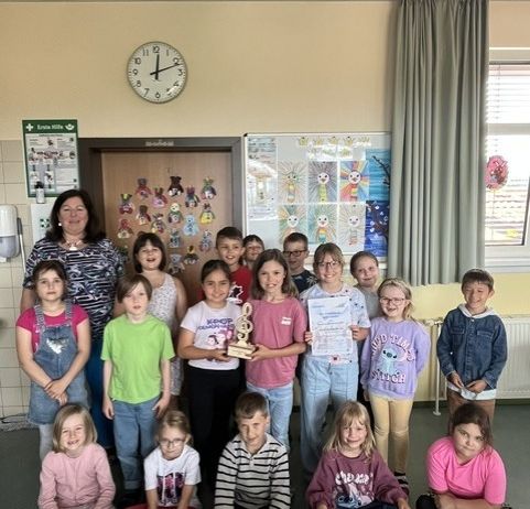 Eine Gruppe von Kindern und einer erwachsenen Frau posiert für ein Foto in einem Klassenzimmer. Sie halten einen Pokal und Urkunden. Das Klassenzimmer hat eine Uhr, eine Tafel und einen Vorhang.