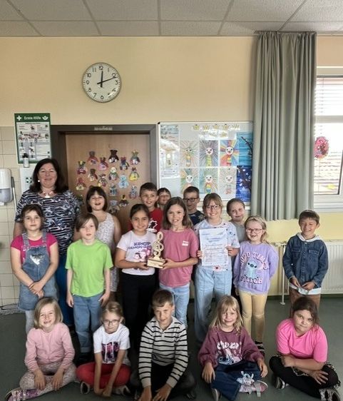 Eine Gruppe von Kindern und einer erwachsenen Frau posiert für ein Foto in einem Klassenzimmer. Sie halten einen Pokal und Urkunden. Das Klassenzimmer hat eine Uhr, eine Tafel und einen Vorhang.
