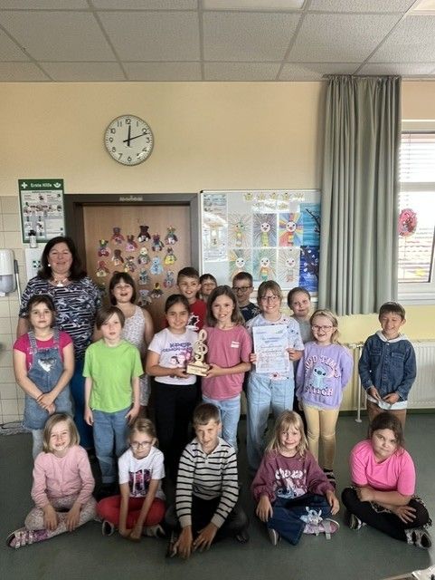Eine Gruppe von Kindern und einer Frau posiert für ein Foto im Klassenzimmer. Sie halten Urkunden und einen Pokal. Das Klassenzimmer hat eine Uhr, einen Aushang und einen Heizkörper.