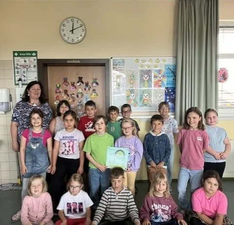Eine Gruppe von Kindern und einer erwachsenen Frau posiert für ein Foto in einem Klassenzimmer. Die Kinder tragen Freizeitkleidung und einige halten Urkunden. Hinter ihnen befindet sich eine Wand mit einer Uhr, einem Whiteboard und einem Vorhang.