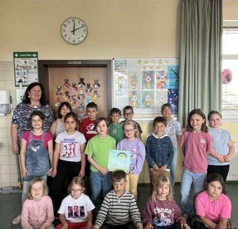 Eine Gruppe von Kindern und einer erwachsenen Frau posiert für ein Foto in einem Klassenzimmer. Die Kinder tragen Freizeitkleidung und einige halten Urkunden. Hinter ihnen befindet sich eine Wand mit einer Uhr, einem Whiteboard und einem Vorhang.