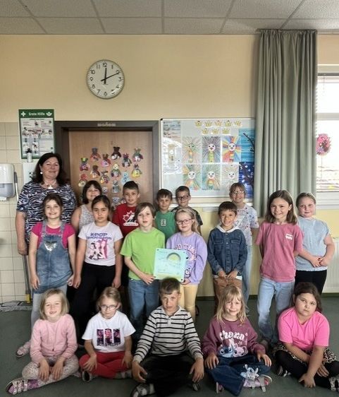 Eine Gruppe von Kindern und einer erwachsenen Frau posiert für ein Foto in einem Klassenzimmer. Die Kinder tragen Freizeitkleidung und einige halten Urkunden. Hinter ihnen befindet sich eine Wand mit einer Uhr, einem Whiteboard und einem Vorhang.