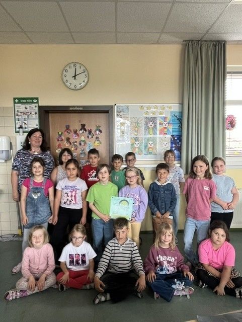 Eine Gruppe von Kindern und ihrer Lehrerin posiert für ein Foto im Klassenzimmer. Die Kinder sitzen auf dem Boden, und einige stehen. Die Lehrerin steht neben ihnen. Hinter ihnen ist eine Wand mit einer Uhr, einem Whiteboard und einer Bulletin-Board.