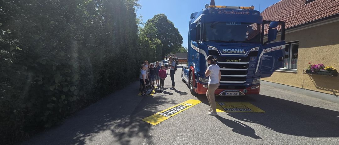 Eine Gruppe von Kindern steht am Straßenrand und beobachtet einen großen blauen Scania-Lkw mit offener Tür. Der Lkw ist in der Nähe eines Gebäudes mit rotem Dach geparkt.