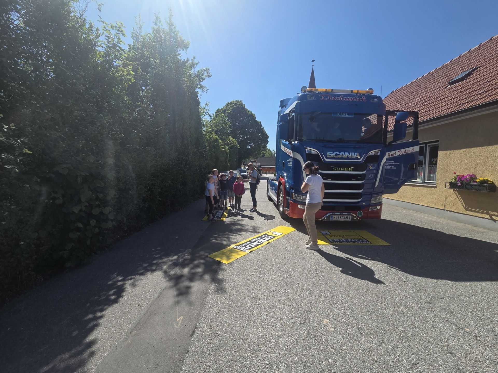 Eine Gruppe von Kindern und Erwachsenen steht am Straßenrand und beobachtet einen blauen Scania-Lkw mit offener Tür. Der Lkw ist mit gelben Markierungen am Straßenrand geparkt.