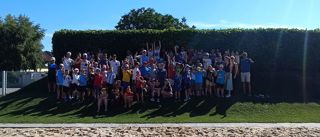 Eine große Gruppe von Menschen, wahrscheinlich ein Sportteam, posiert für ein Foto auf einem Rasenfeld mit einem sandigen Bereich und einem Hecke im Hintergrund.